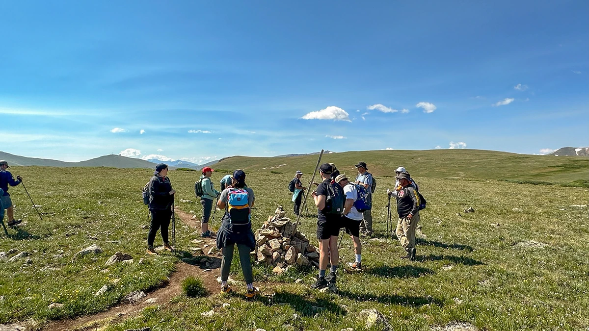 Group of Cairn participants pausing for a break and looking at the grassy rolling hill scenery and standing around a rock trail marker.