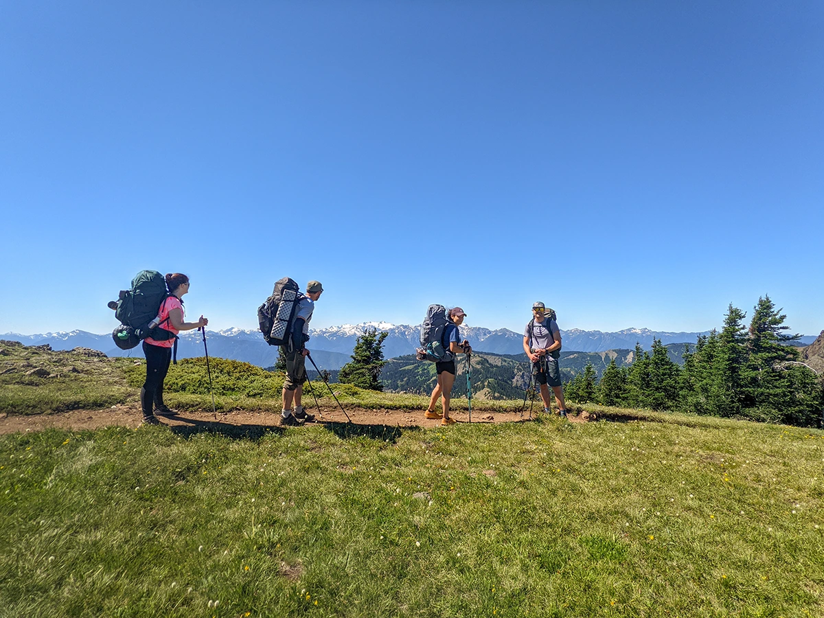 Cairn backpacker group of four stopping for a quick break and standing on the trail with scenic snowcapped mountains in the distance.