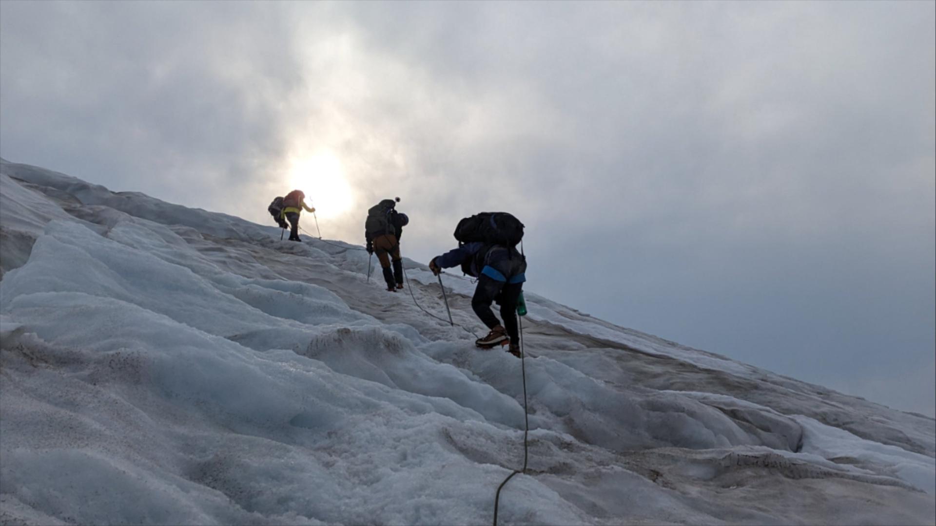 Three climbers scale a steep slope of snow and ice as dawn breaks through parting clouds.