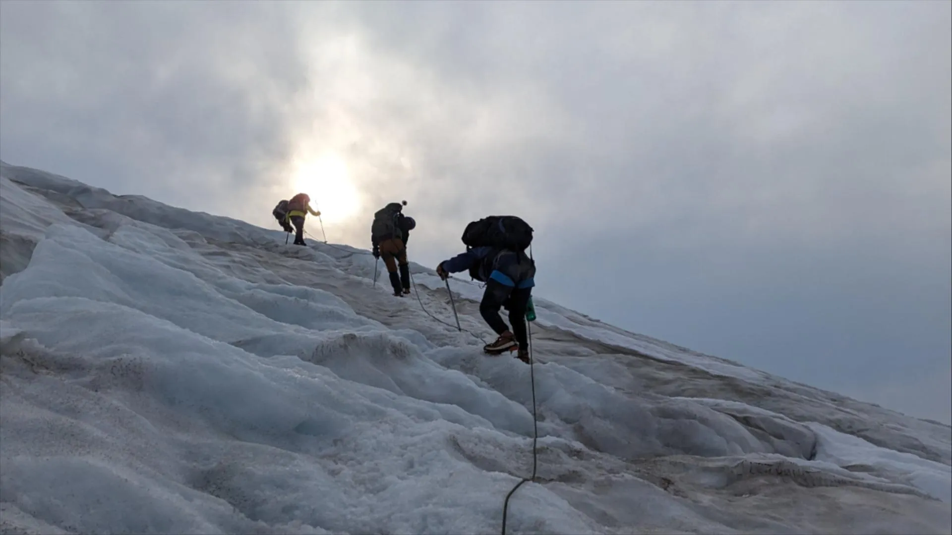 Four climbers, roped together, scale a steep slope of snow and ice as dawn breaks through parting clouds.