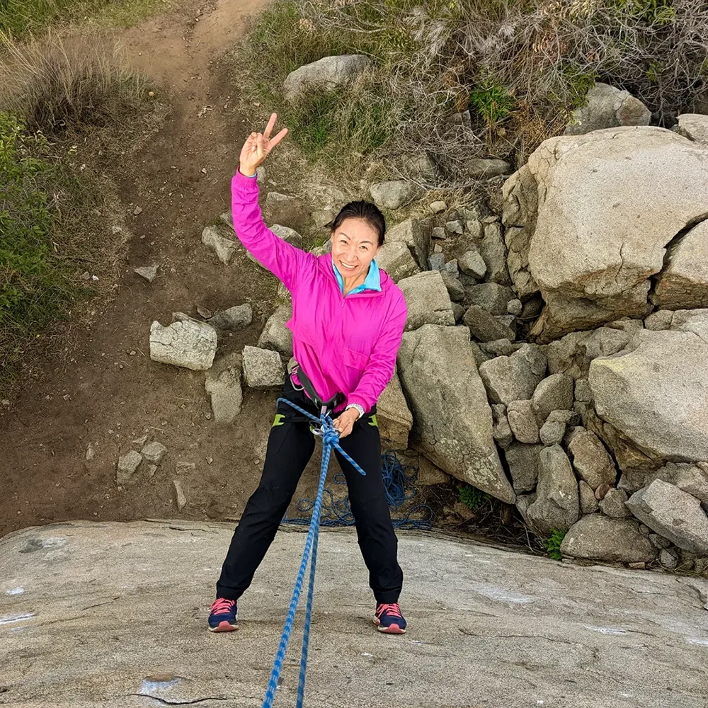 A Cairn Leadership client pushing her boundaries by learning to rock climb for the very first time looking at the camera and smiling while rappelling down a rock face.