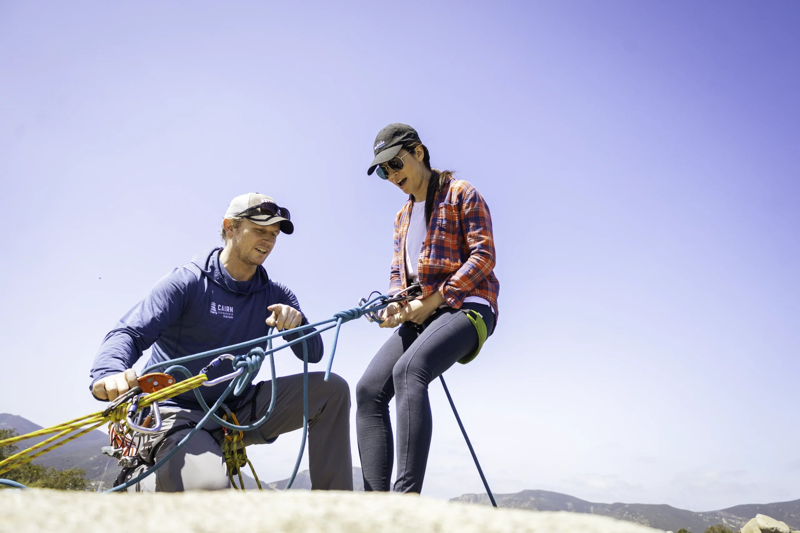 At the top of a rock cliff with a sunny blue sky in the background, a guide prepares a client to rappel down ropes anchored to the cliff.
